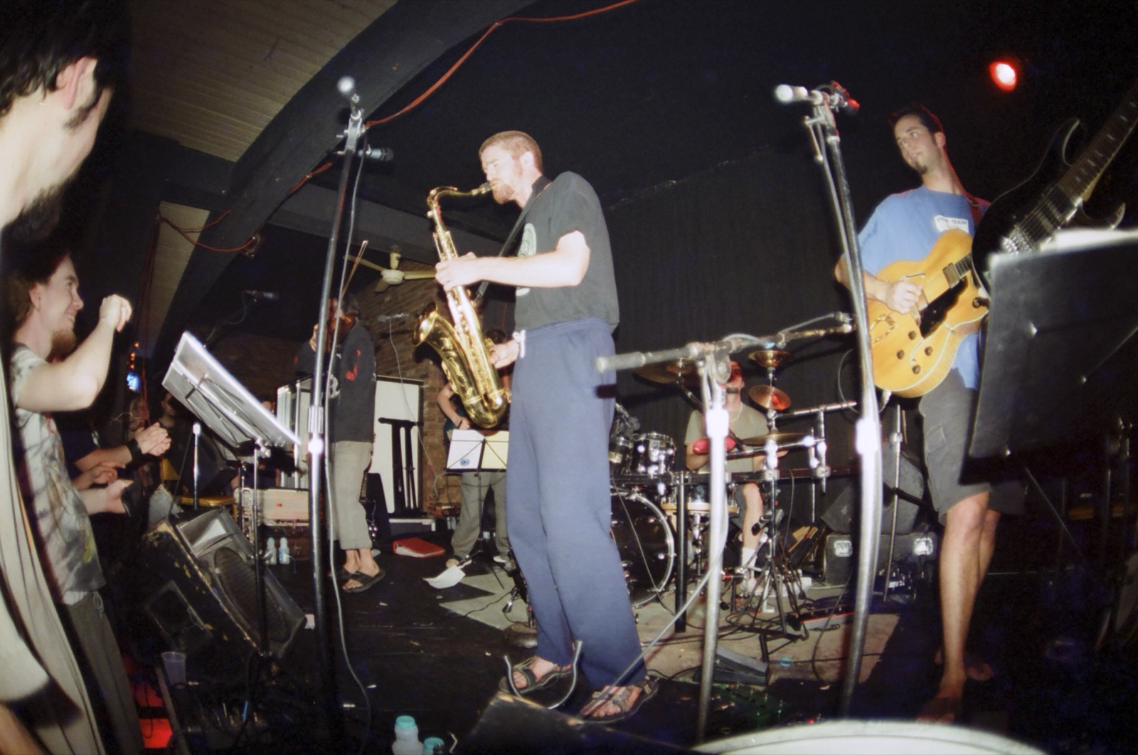 A wide fisheye view of a small club stage with a saxophonist, guitarist and drummer as fans cheer at the edge.